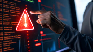Hand pointing at a glowing red warning triangle with an exclamation mark on a computer screen displaying code, suggesting a cybersecurity alert or system error.